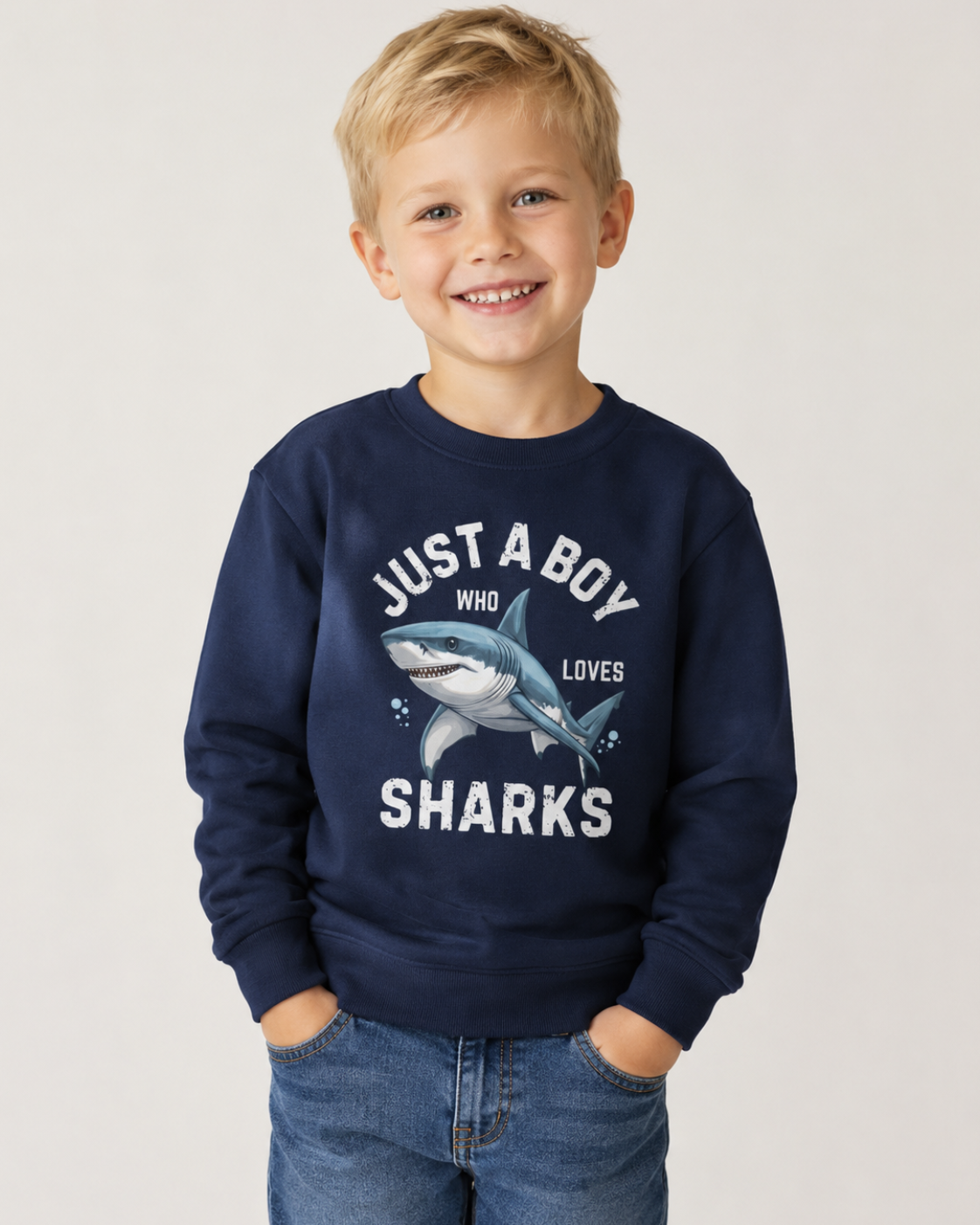A young boy with blonde hair smiling, wearing a navy blue crewneck sweatshirt with a shark graphic and the text 'JUST A BOY WHO LOVES SHARKS', paired with blue jeans, against a plain light grey studio background.