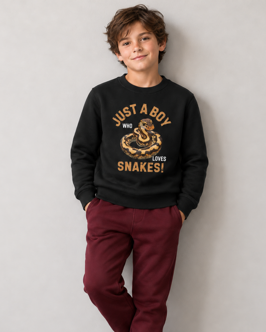 A young boy wearing a black crewneck sweatshirt with a 'Just a boy who loves snakes!' graphic and burgundy pants, standing against a light gray wall.