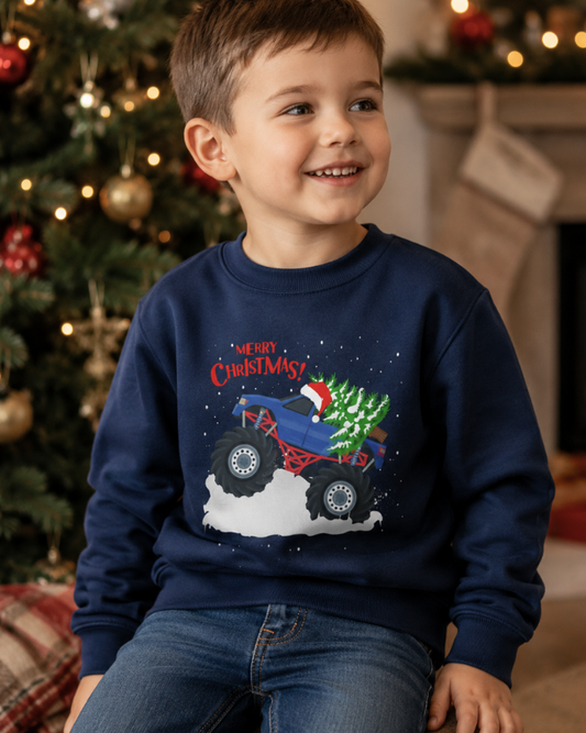A young boy wearing a navy blue crewneck sweatshirt with a 'Merry Christmas' monster truck graphic, sitting in front of a decorated Christmas tree and fireplace.