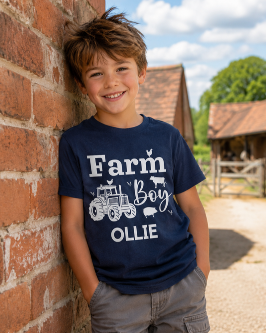 A smiling young boy with brown hair leans against a red brick cottage wall outdoors on a farm, wearing a navy blue graphic t-shirt that reads 'Farm Boy' with the name OLLIE featuring a tractor and farm animal illustrations, paired with grey cargo trousers.