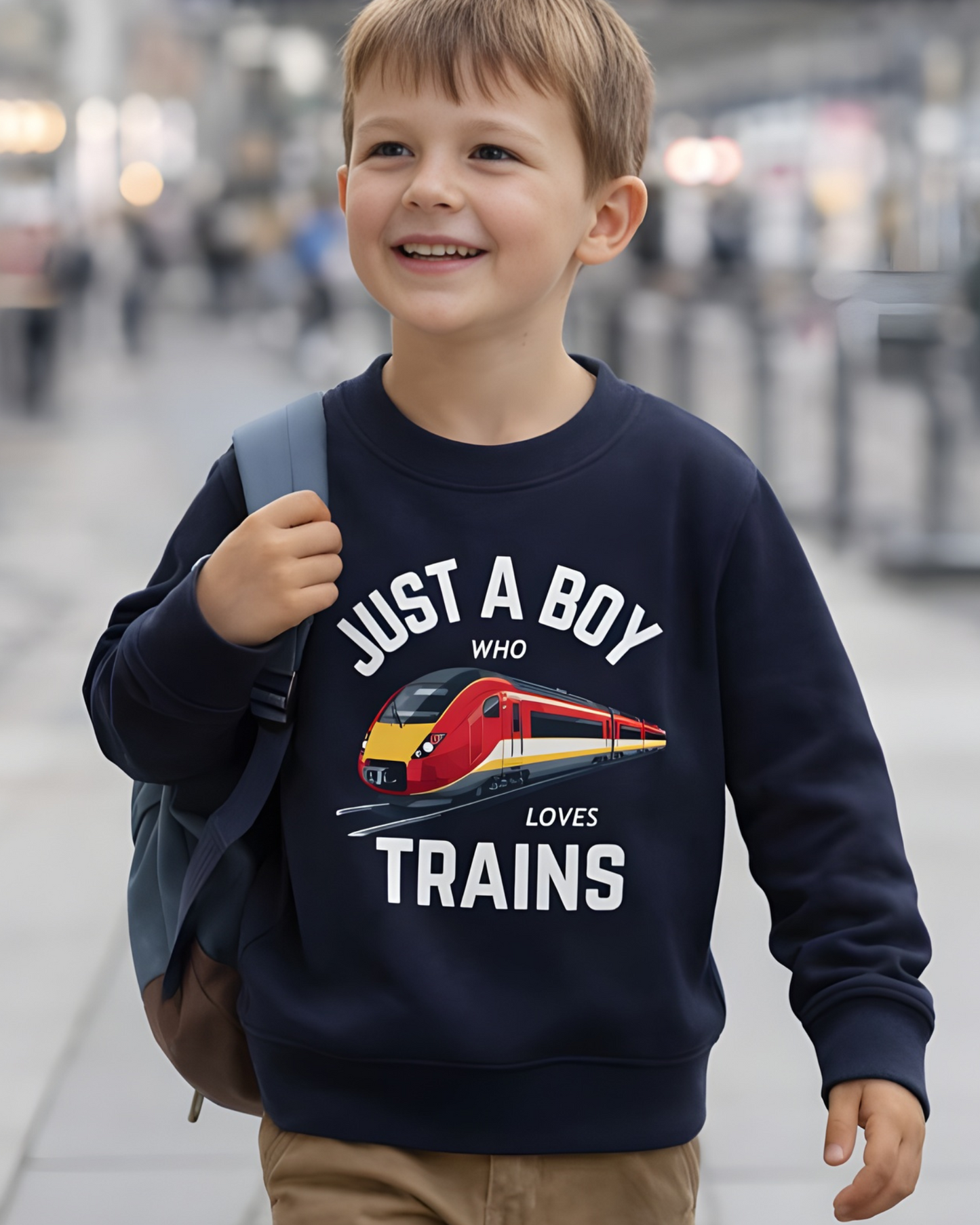 Young boy wearing navy “Just a Boy Who Loves Trains” sweatshirt with red train graphic, walking through a modern train station with backpack.