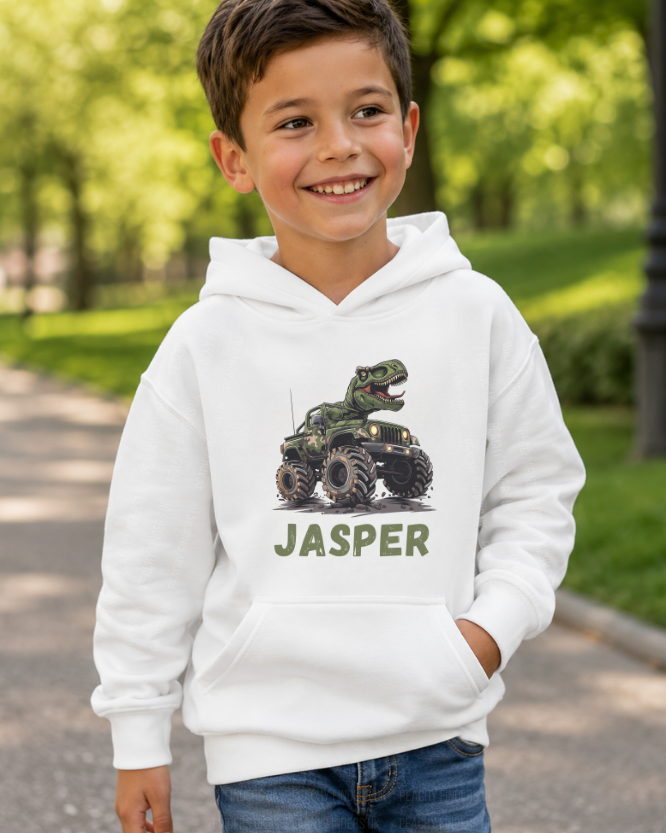 Smiling boy wearing white personalised hoodie with T-Rex riding camo monster truck and name Jasper printed on front - park background.