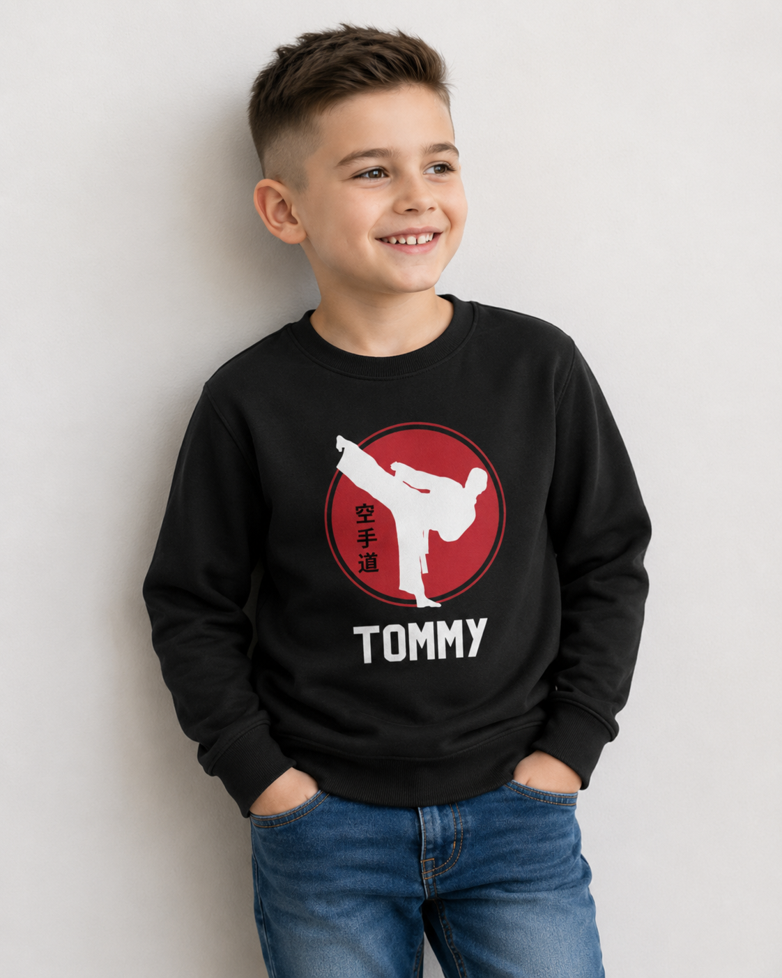 Boy wearing a personalised black karate sweatshirt with custom name, red circle design, white karate kick silhouette and Japanese kanji text, styled with jeans in a studio setting.