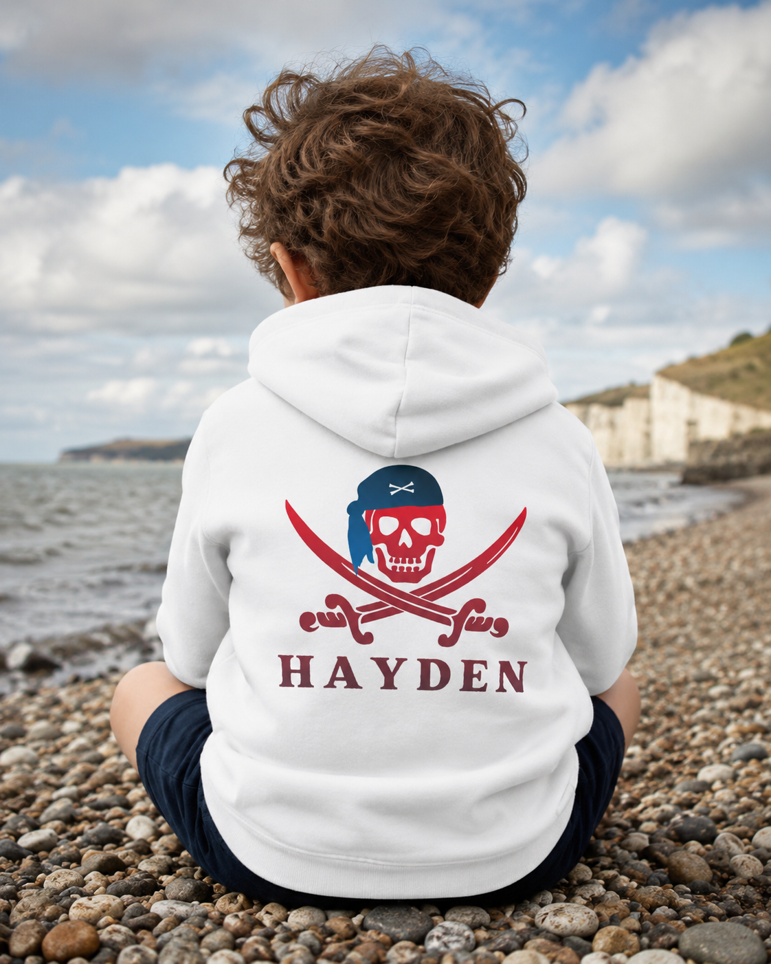 A little boy seen from behind sitting on a pebble beach wearing a white hoodie with a pirate skull and crossed swords graphic on the back, above the name 'HAYDEN' in red. The background shows the ocean, sky, and shoreline.