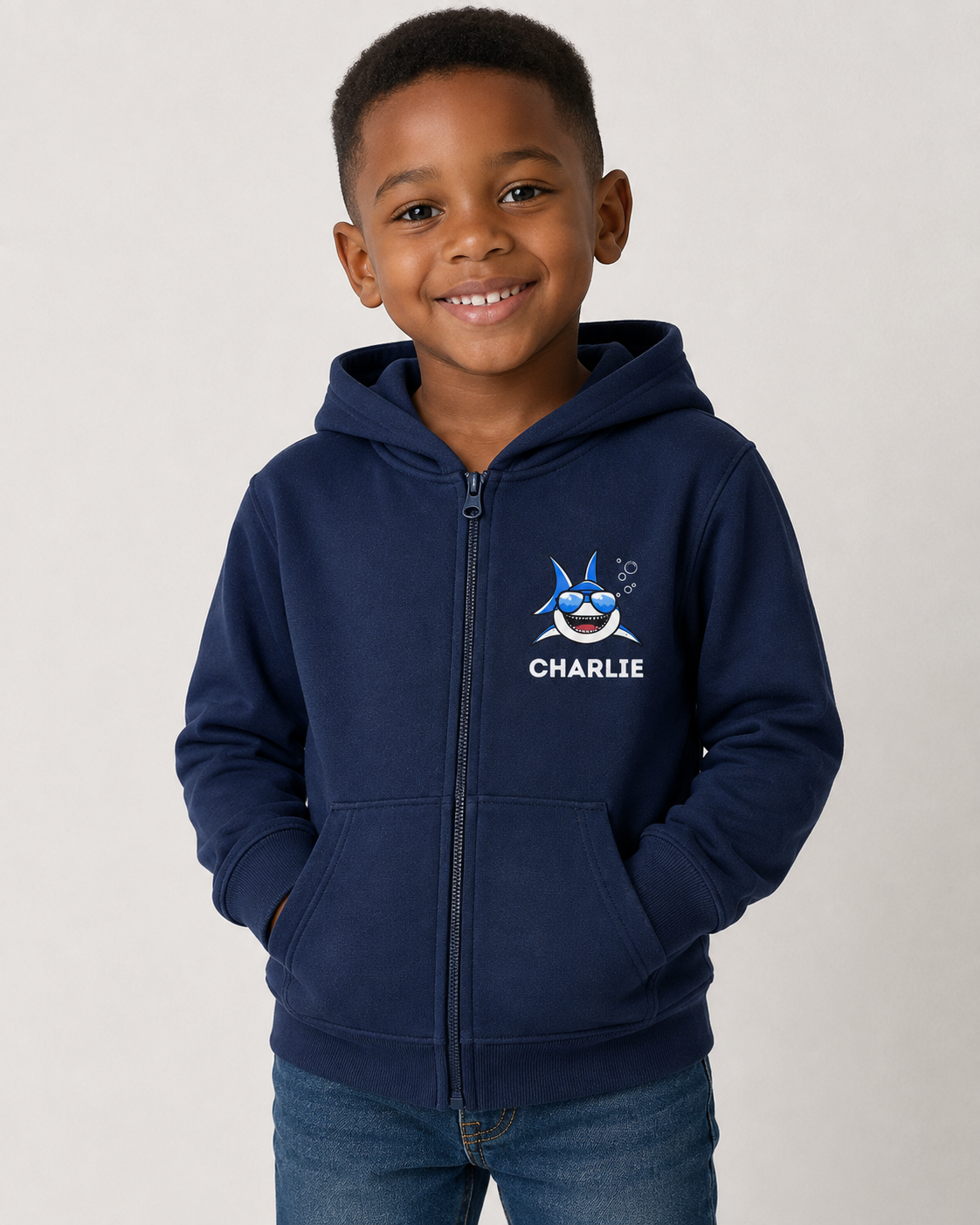 A smiling young boy wearing a navy blue zip-up hoodie with a cartoon shark graphic and the name 'CHARLIE' printed on the chest, paired with blue jeans, against a plain light grey background.