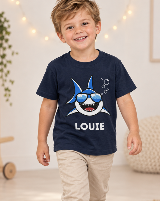 Four-year-old boy wearing a personalised navy T-shirt featuring a smiling shark in sunglasses with his name underneath.
