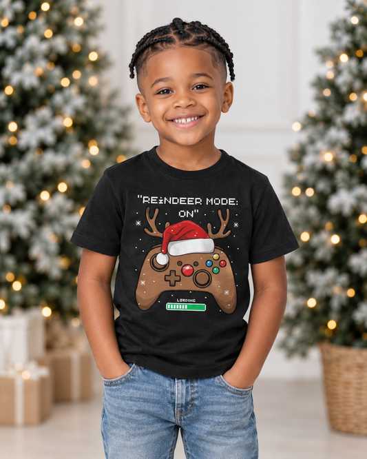 Smiling six-year-old boy wearing a black Reindeer Mode On gaming Christmas T-shirt with jeans beside festive trees.