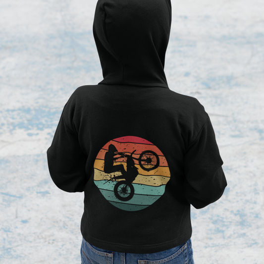 A little boy wearing a black hoodie with a back printed graphic of a person on a dirt bike against a sunset background.