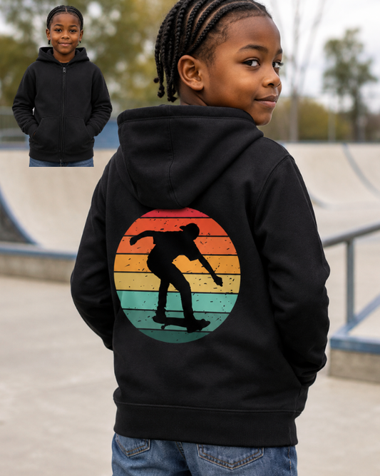 A child wearing a black hoodie with a large retro sunset graphic and skateboarder silhouette on the back, shown outdoors at a skatepark. A small inset in the upper left shows the child from the front wearing the hoodie.