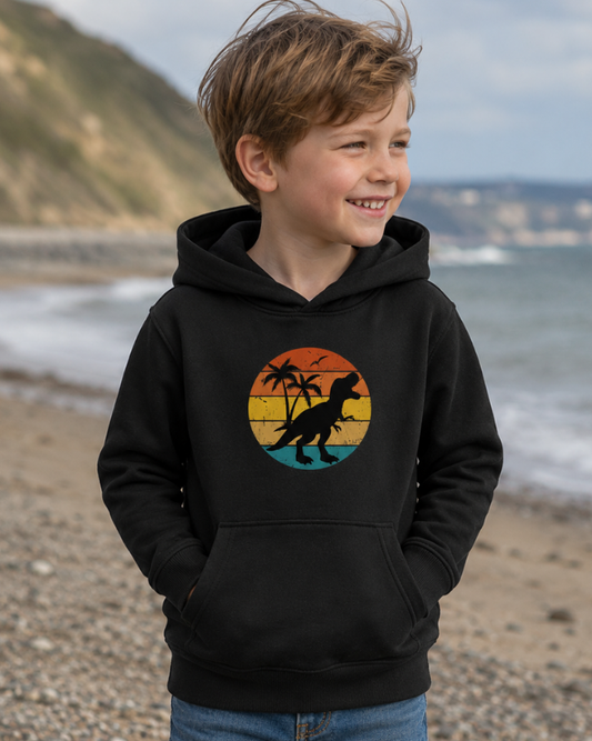Smiling boy wearing a black T-rex sunset graphic hoodie on a breezy beach, shown in a natural lifestyle photo for kids dinosaur clothing.
