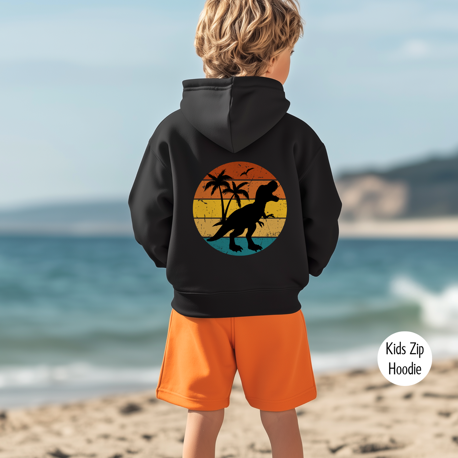 Boy wearing black kids zip hoodie with retro sunset T-Rex back print at the beach