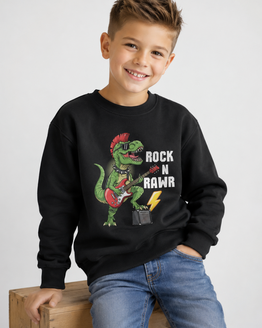 A smiling young boy wearing a black crewneck sweatshirt with a colourful graphic of a green dinosaur playing a red guitar and the text 'ROCK N RAWR', paired with blue jeans, sitting on a wooden crate against a light grey background.