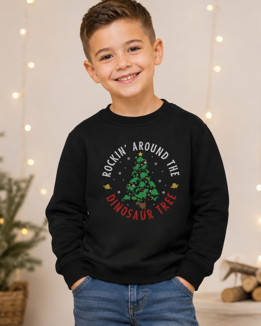 A young boy smiling and wearing a black crewneck sweatshirt with a Christmas-themed graphic that reads 'ROCKIN' AROUND THE DINOSAUR TREE' in white and red text around a green Christmas tree. The background is a warm indoor setting with soft string lights.