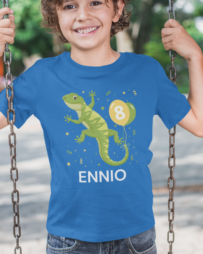 A smiling boy wearing a bright blue custom graphic t-shirt on a swing. The shirt features a green cartoon gecko, a yellow balloon with the number 8, and the name 'ENNIO' printed in white.