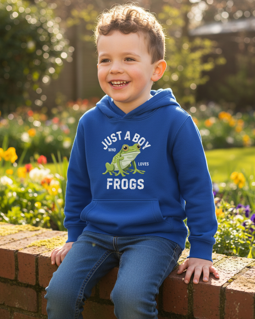 A young boy smiling while sitting on a brick wall in a garden, wearing a royal blue hoodie with a green frog graphic and the text 'JUST A BOY WHO LOVES FROGS'.