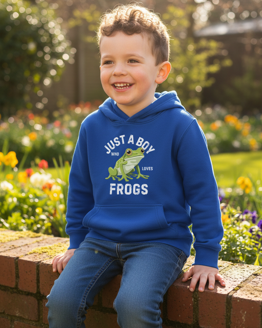 A young boy smiling while sitting on a brick wall in a garden, wearing a royal blue hoodie with a green frog graphic and the text 'JUST A BOY WHO LOVES FROGS'.