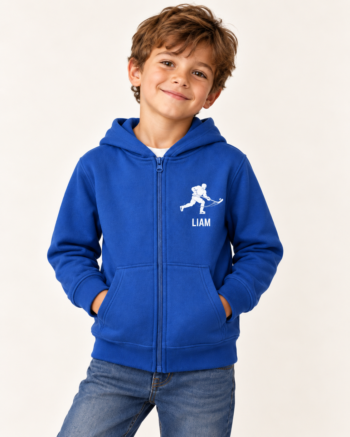 A young boy smiling while wearing a royal blue zip-up hoodie with a white hockey player graphic and the name 'LIAM' printed on the chest, paired with blue jeans against a plain off-white background.