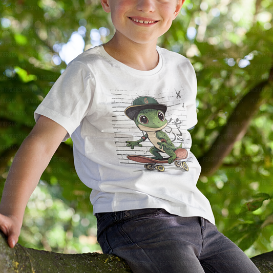 A smiling child sits on a tree branch, wearing a white t-shirt with a cartoon graphic of a green gecko skateboarding in front of a graffiti-style brick wall. The gecko wears a backwards baseball cap with a red "G".