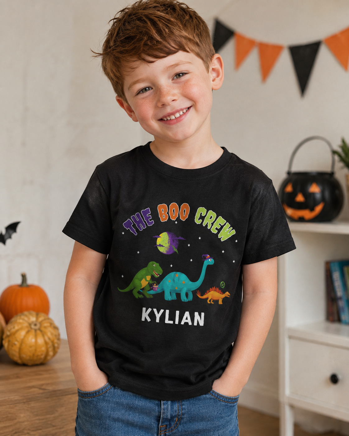 A young boy wearing a black graphic t-shirt with 'THE BOO CREW' and the name 'KYLIAN' printed on the front, styled in a cozy, decorated home setting with pumpkins and Halloween bunting.
