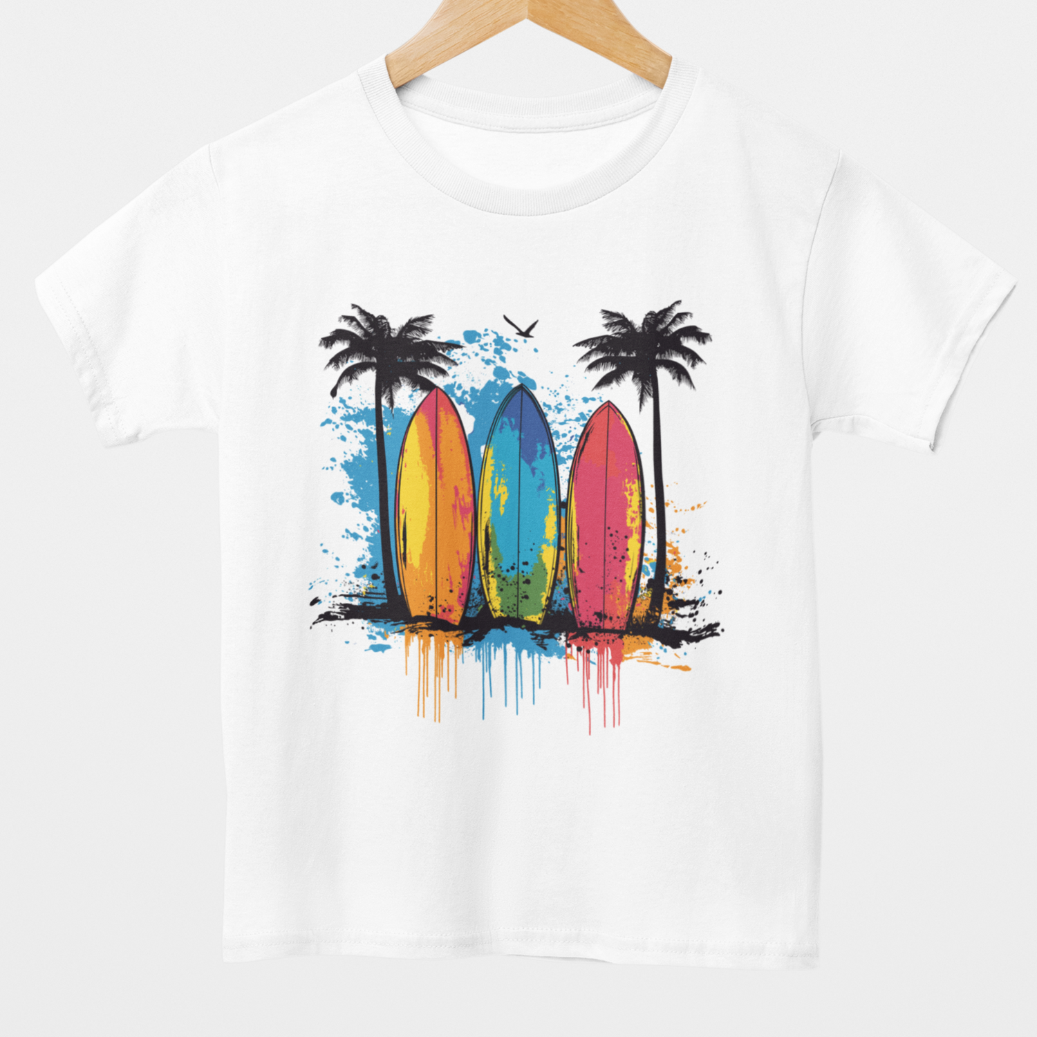A kids white short-sleeved t-shirt hanging on a wooden hanger. The t-shirt features a vibrant graphic of three surfboards (yellow, blue, and pink) leaning against each other on a sandy surface with splatters of blue, orange, and black paint. Two black palm trees stand on either side of the surfboards, and a black bird is flying overhead.