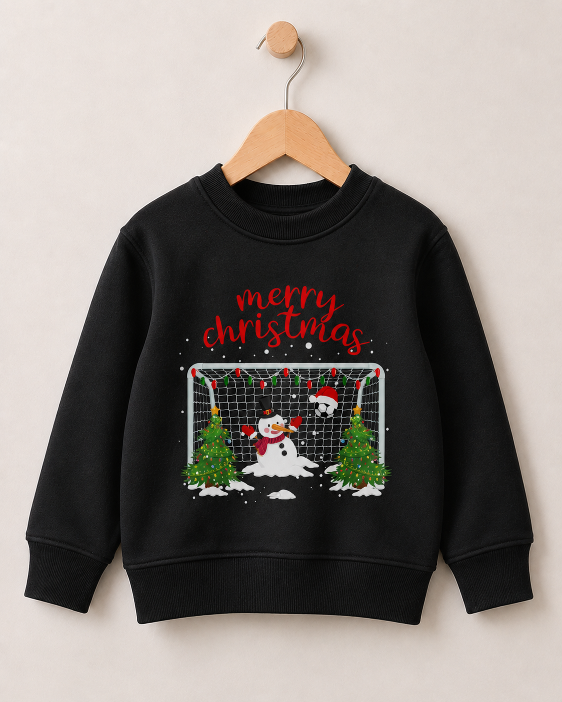 Children's black crewneck sweatshirt hanging on a wooden hanger against a light beige wall. The sweatshirt features a festive Christmas graphic with a snowman, soccer ball, Christmas trees, and the words 'merry christmas' in red script.