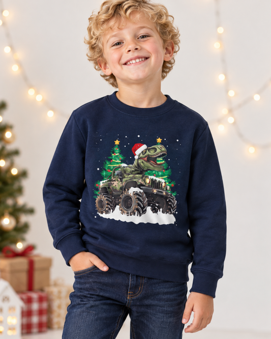 A smiling young boy with curly blonde hair wearing a navy crewneck sweatshirt with a festive graphic of a green dinosaur driving a military-style vehicle in front of Christmas trees and snow, paired with dark blue jeans. The background is softly lit with warm string lights and blurred holiday decorations.