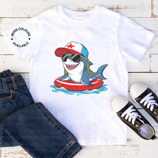 White t-shirt with a cartoon shark design wearing sunglasses, a cap, and a life buoy, paired with jeans and sneakers on a wooden surface.