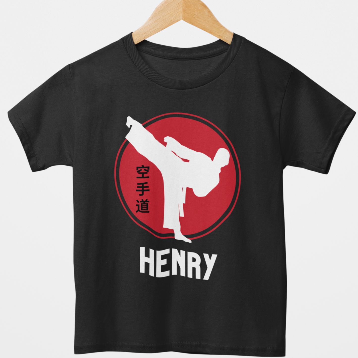 Boys black personalised karate T-shirt featuring a white martial artist silhouette performing a high kick inside a red circle with Japanese kanji text, and custom name, styled on a wooden hanger.