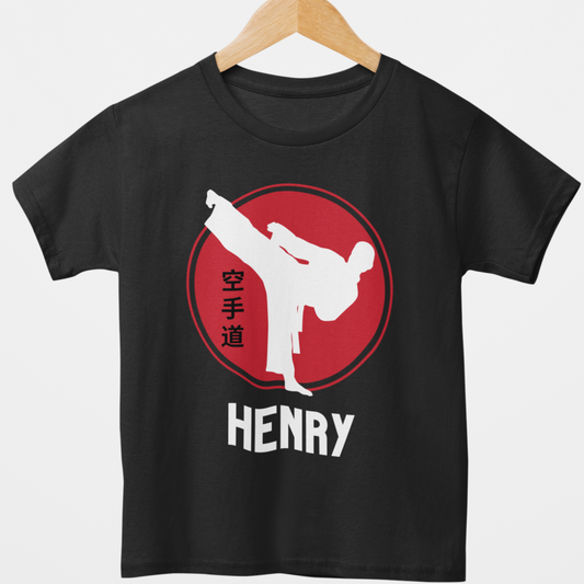 Boys black personalised karate T-shirt featuring a white martial artist silhouette performing a high kick inside a red circle with Japanese kanji text, and custom name, styled on a wooden hanger.