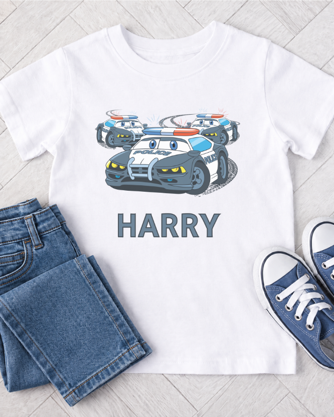 Kids white t-shirt with cartoon police cars and 'HARRY' text, paired with blue jeans and pumps on a light wooden floor.
