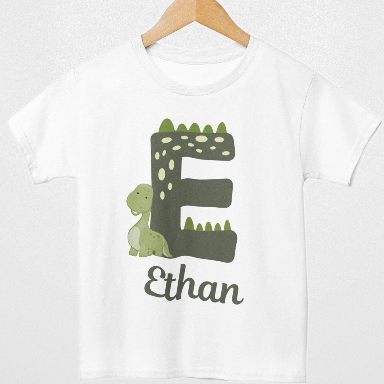 White kids T-shirt on a wooden hanger with large green dinosaur-themed letter E, small green dinosaur and personalised name Ethan printed underneath.