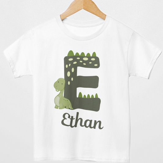 White kids T-shirt on a wooden hanger with large green dinosaur-themed letter E, small green dinosaur and personalised name Ethan printed underneath.
