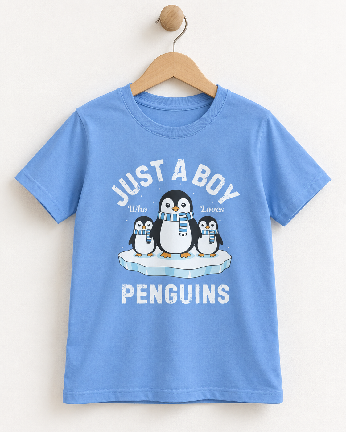Light blue boys’ T-shirt on a wooden hanger, featuring three penguins in striped scarves on an ice floe and “Just a Boy Who Loves Penguins” lettering.