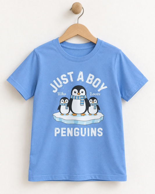 Light blue boys’ T-shirt on a wooden hanger, featuring three penguins in striped scarves on an ice floe and “Just a Boy Who Loves Penguins” lettering.