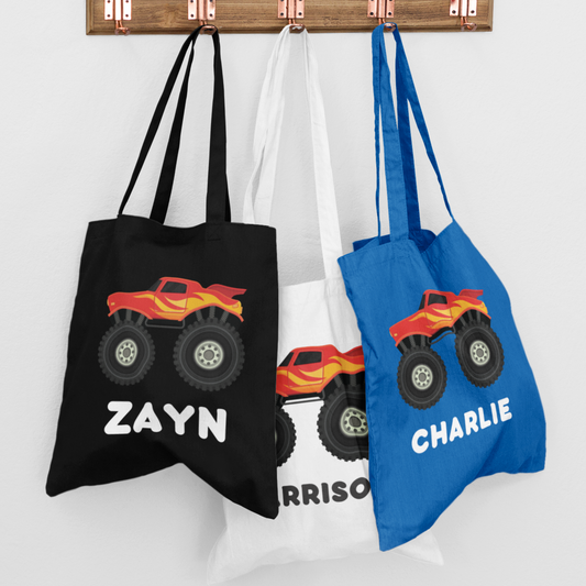 Three colourful boys tote bags featuring a red and yellow monster truck design are hanging on wall hooks. Each bag is personalised with a custom name.