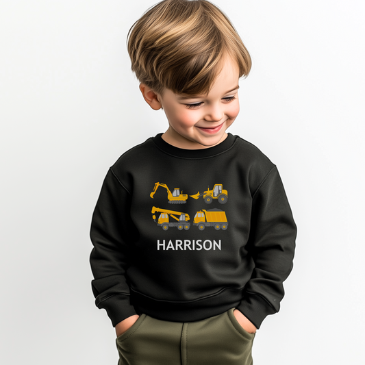 A young boy modelling a black sweatshirt with yellow construction vehicles - excavator, bulldozer, tractor, crane truck and tipper -custom name “Harrison” printed underneath in a white font.