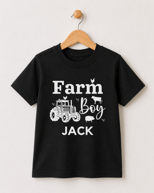 A black short-sleeve kids t-shirt hanging on a wooden hanger against a light beige wall. The shirt features a white graphic print that reads 'Farm Boy JACK' with illustrations of a tractor, a cow, a pig, and chickens.