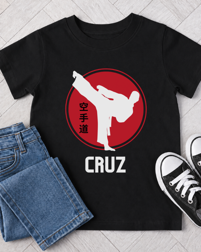 A black short-sleeve T-shirt laid flat on a light wood floor, styled with folded blue jeans and black sneakers. The shirt has a large red circular graphic with a white karate figure kicking with the name 'CRUZ' printed below.