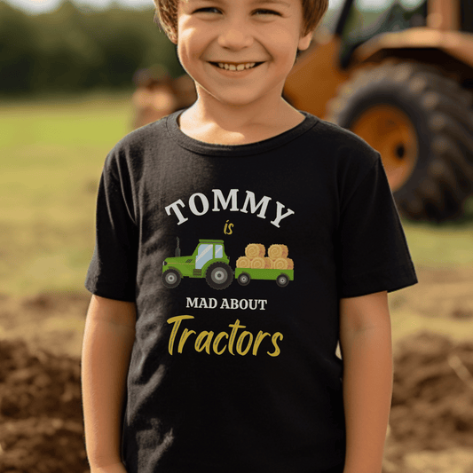 Young boy standing in a field wearing a black short sleeved t-shirt with a printed green tractor and trailer with bales of hay