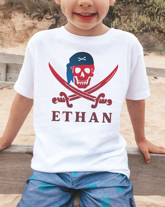 Boy wearing white personalised pirate skull and swords t-shirt with name Ethan, beach background