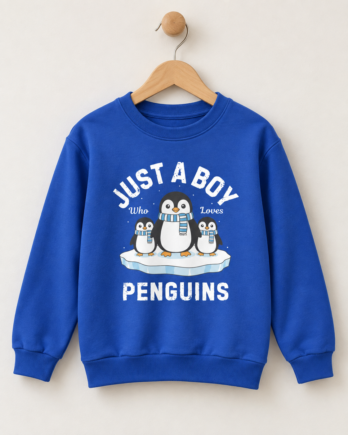 Royal blue boys sweatshirt on a wooden hanger and wall hook, featuring three penguins on an ice floe and distressed white lettering.