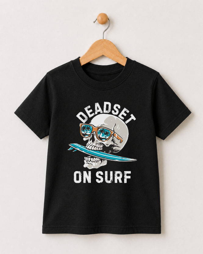 A black short-sleeve t-shirt hanging on a wooden hanger against a light beige wall. The shirt features a graphic print of a skull wearing orange sunglasses and holding a blue surfboard, with the text 'DEADSET ON SURF' in white.