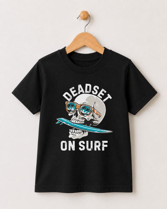 A black short-sleeve t-shirt hanging on a wooden hanger against a light beige wall. The shirt features a graphic print of a skull wearing orange sunglasses and holding a blue surfboard, with the text 'DEADSET ON SURF' in white.