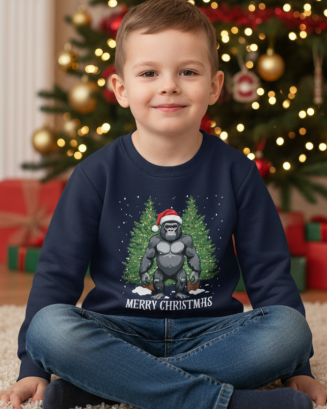 A young boy sitting cross-legged in front of a decorated Christmas tree, wearing a navy blue sweatshirt with a graphic of a gorilla in a Santa hat and the text 'MERRY CHRISTMAS'.
