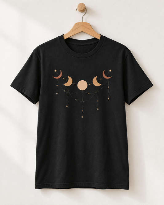 A black short-sleeve crewneck t-shirt hanging on a wooden hanger against a light beige wall. The shirt features a celestial graphic of moon phases and stars in a muted peach and gold colour on the chest.