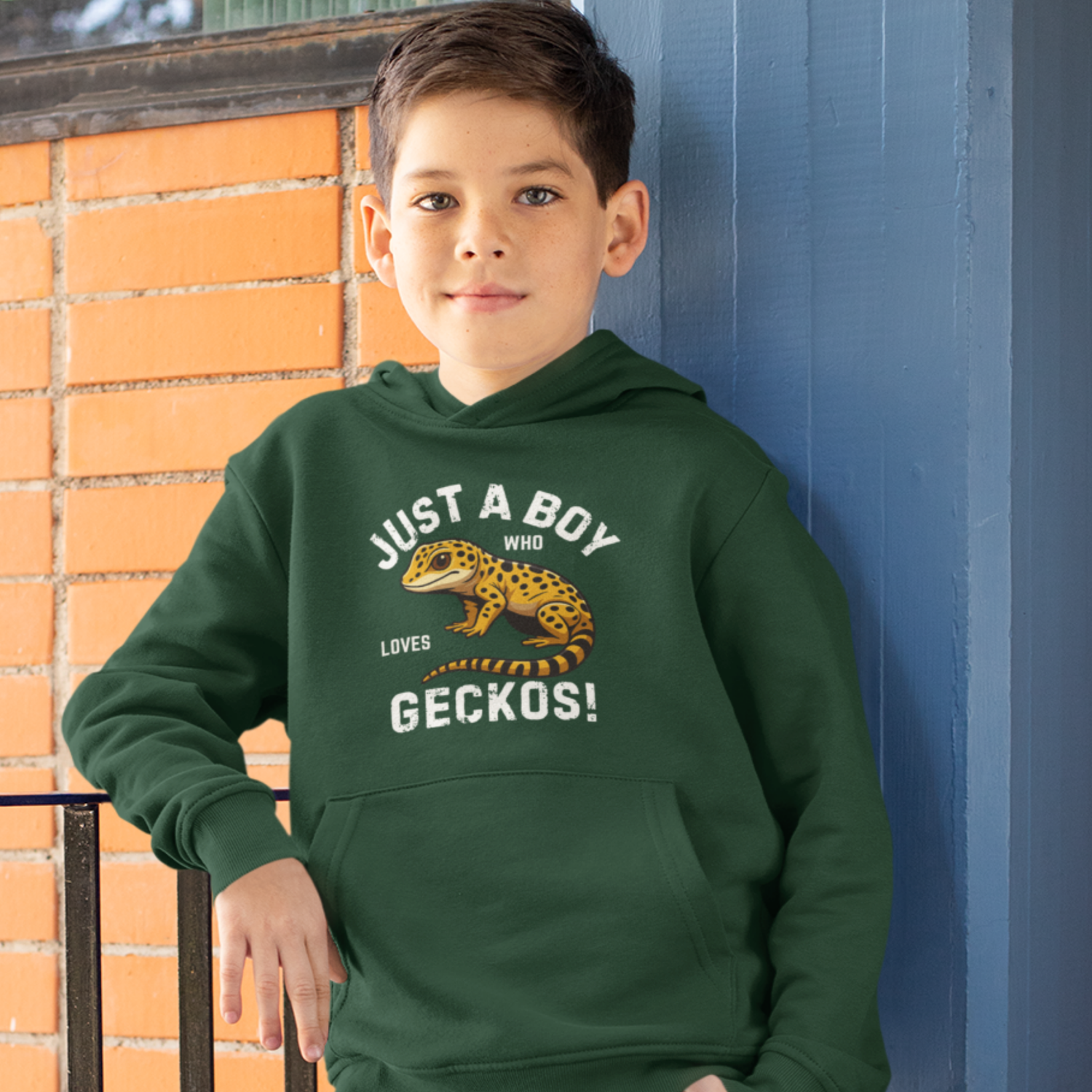 Child wearing a green hoodie with a gecko graphic and text, standing against a blue wall.