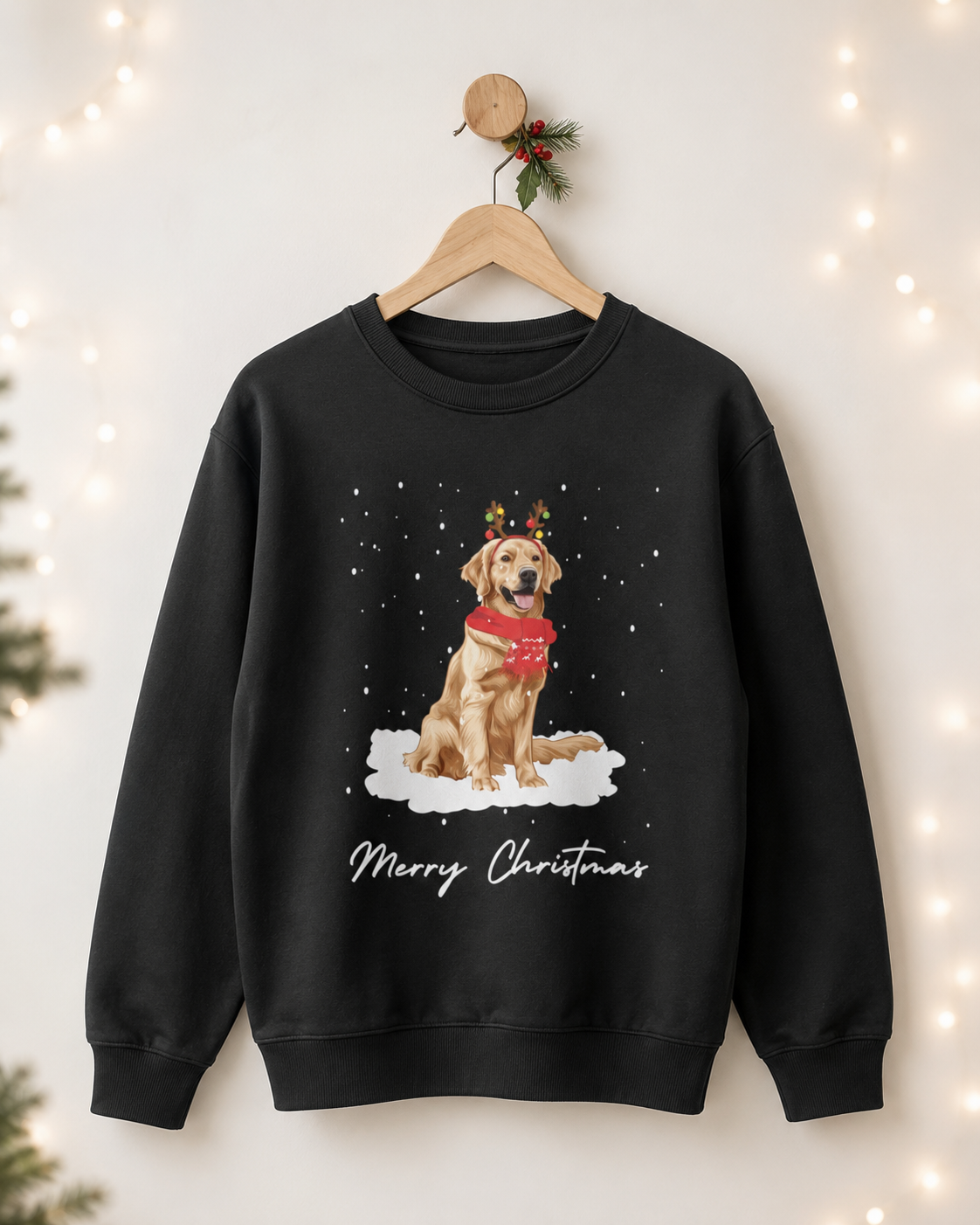 Adults black crewneck sweatshirt hanging on a wooden hanger against a warm, softly lit background with string lights. The sweatshirt features a graphic of a golden retriever wearing a red scarf and reindeer antlers, sitting in a snowy white cloud, with 'Merry Christmas' written below.