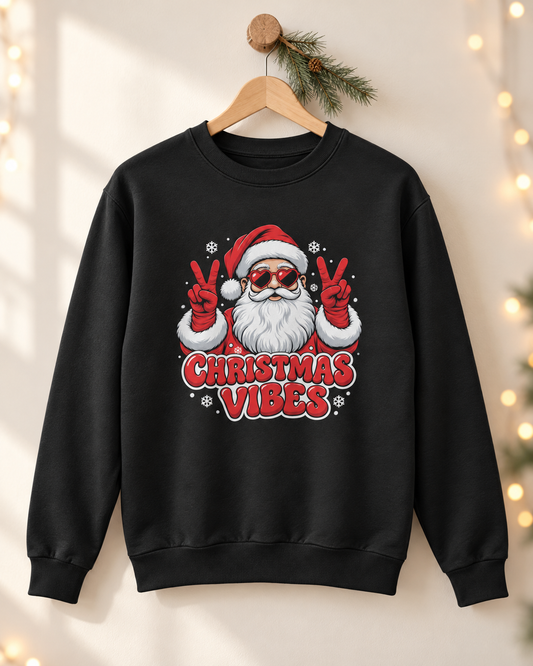 Adults black crewneck sweatshirt hanging on a wooden hanger decorated with a pine branch, featuring a festive graphic of Santa Claus wearing sunglasses and making peace signs with the words 'CHRISTMAS VIBES' in red text below.