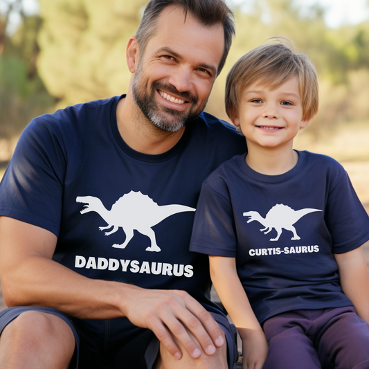 “Dad and son wearing matching navy dinosaur T-shirts, one with ‘DADDYSAURUS’ and the other personalised with ‘CURTIS-SAURUS’, sitting together and smiling outdoors.”