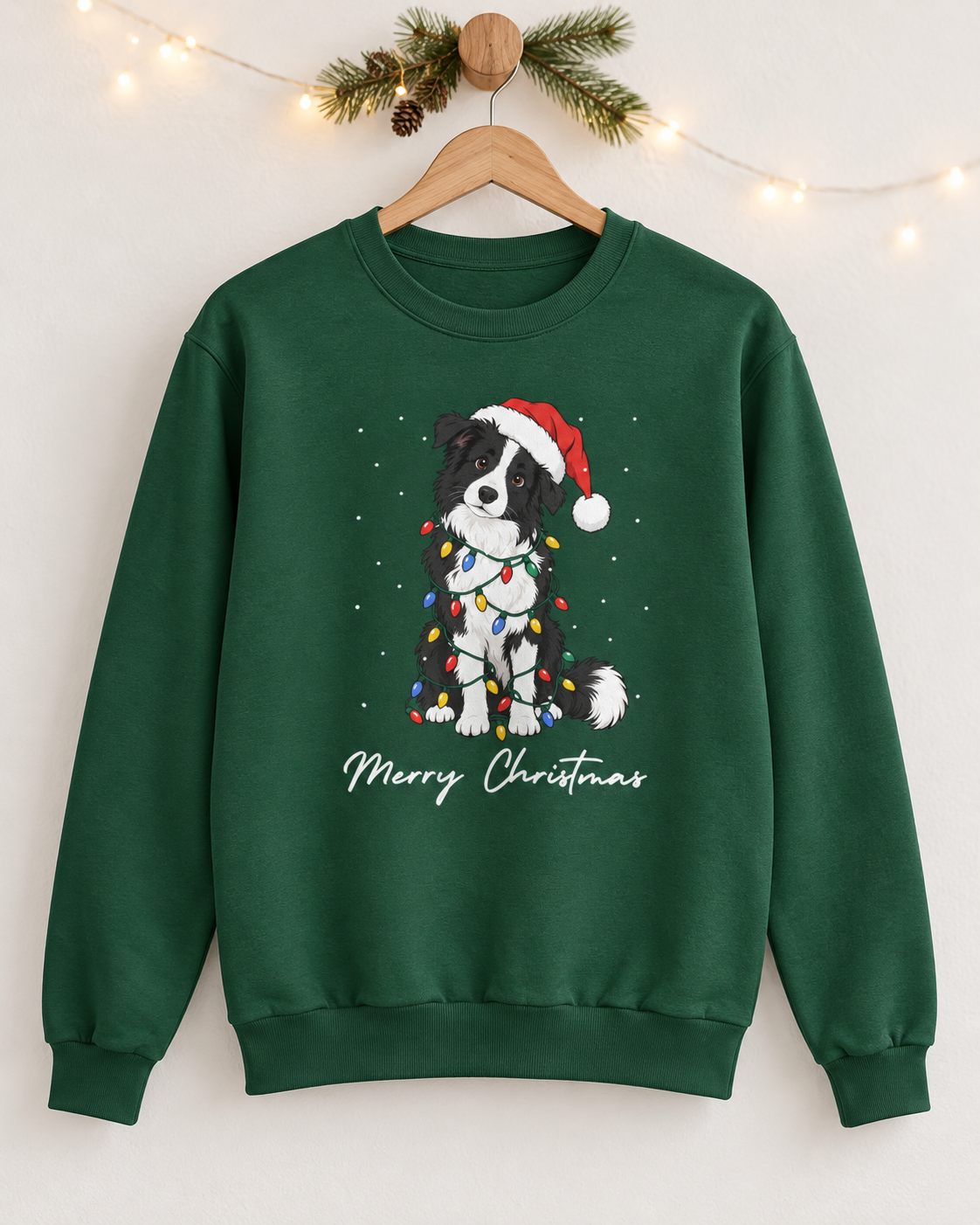 A dark green adult Christmas sweatshirt hanging on a wooden hanger against a neutral wall decorated with pine branches and warm fairy lights, displaying the Border Collie holiday graphic.
